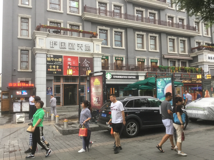 Grey building and storefronts. A few pedestrians walk through the foreground.