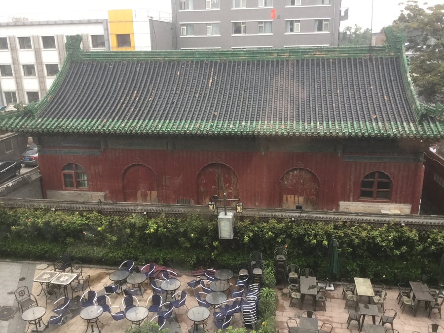 A patio with several café tables in front of a large, worn, red building with a dark roof.