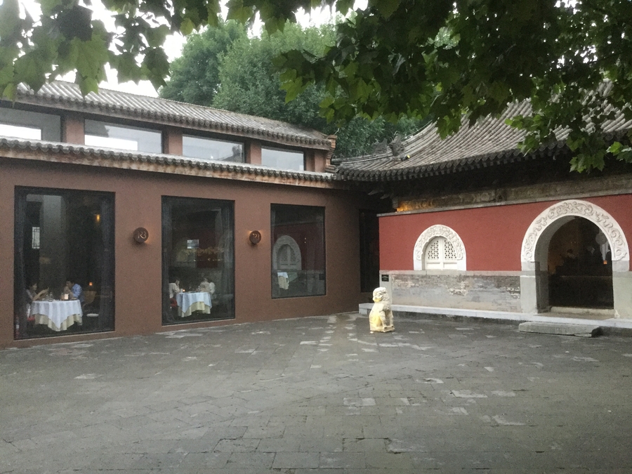 Courtyard with a brown building with windows showing a dining room with patrons and a red building with an elaborate arched entryway leading into a dark interior