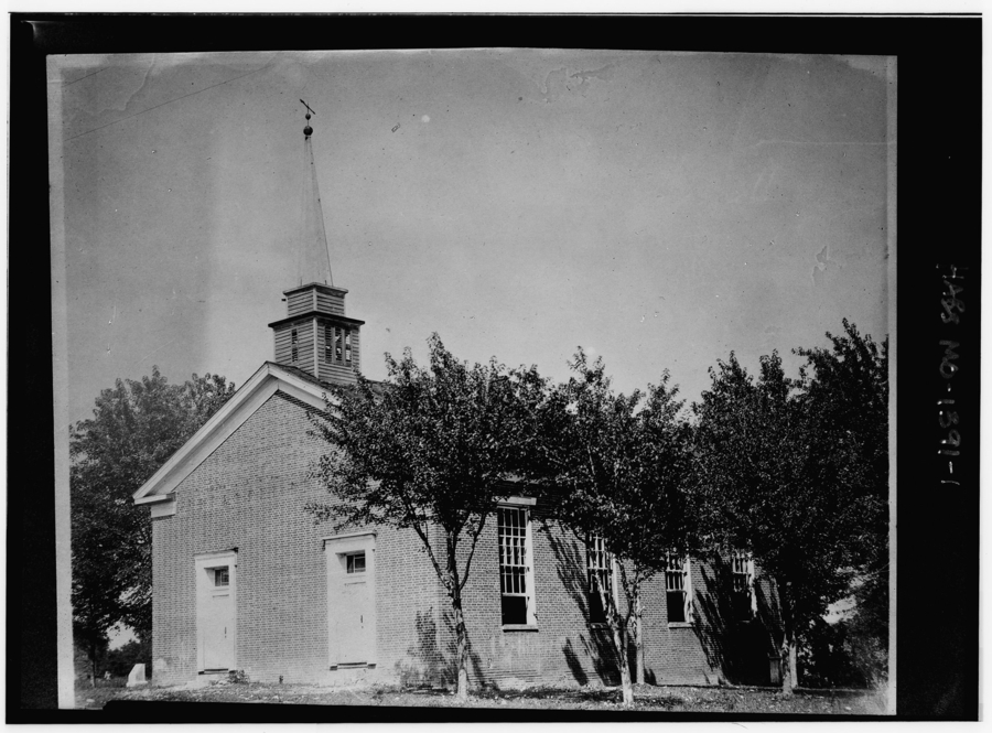 A black and white photo captures a one-story, brick church. The church has a cross-gable roof with a bell tower and steeple. 