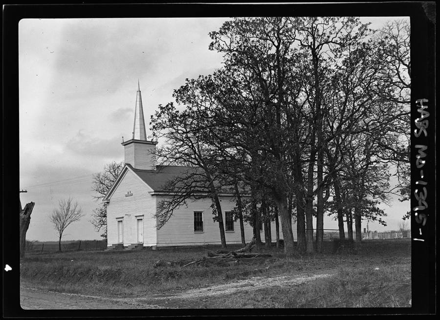 A one-story, gable-ended church stands beside a group of trees in a black and white photo. The sparse landscape includes a small cemetery behind the church. 