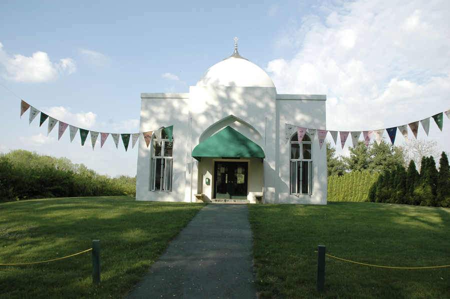A white building has a rectangular base with an onion dome on top and a green awning over the entrance. Banners of triangular flags emanate from the sides of the entrance and are fastened at some distance away from the photo frame.