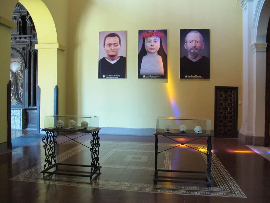 In a museum interior, a case of three skulls and a case of three cast masks are positioned in front of three digital portraits.