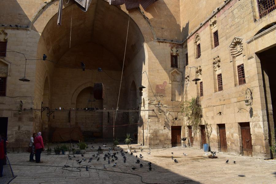 In a view from within the courtyard of a khānqāh, the wood doors of khalwa cells are visible on the ground floor of the building. The windows of living quarters line the walls above them. The courtyard itself has a massive pointed arch on one end.