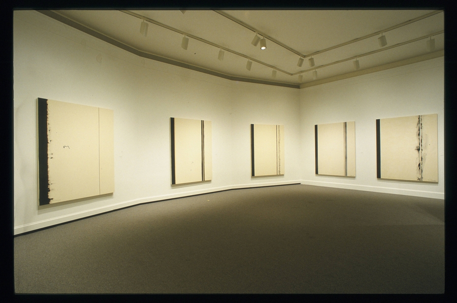A series of large white and black paintings hangs on the walls of a white gallery space. The white canvases are painted with black lines of differing thicknesses. 