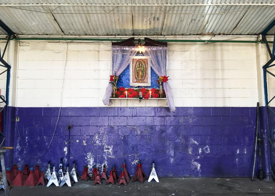 Hanging on a purple and white brick wall, a white frame forms an altar in an industrial workspace. The altar frames an image of Our Lady of Guadalupe against a blue fabric backdrop and behind gauzy pink drapes. Poinsettias line the altar.