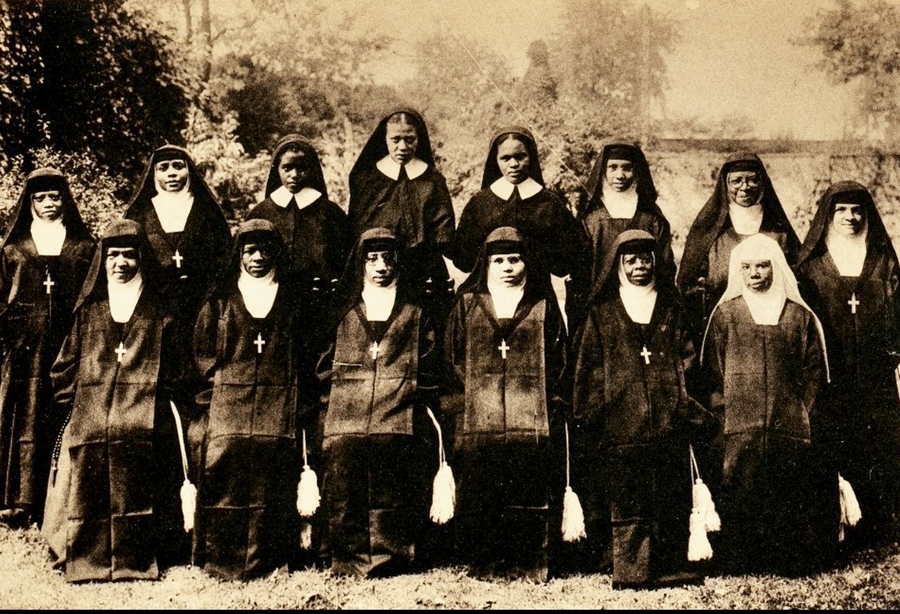 Two rows of magdalens--one kneeling, the other standing behind--are photographed in black habits in an outdoor setting. They are posed on a grassy field in front of a treelike.