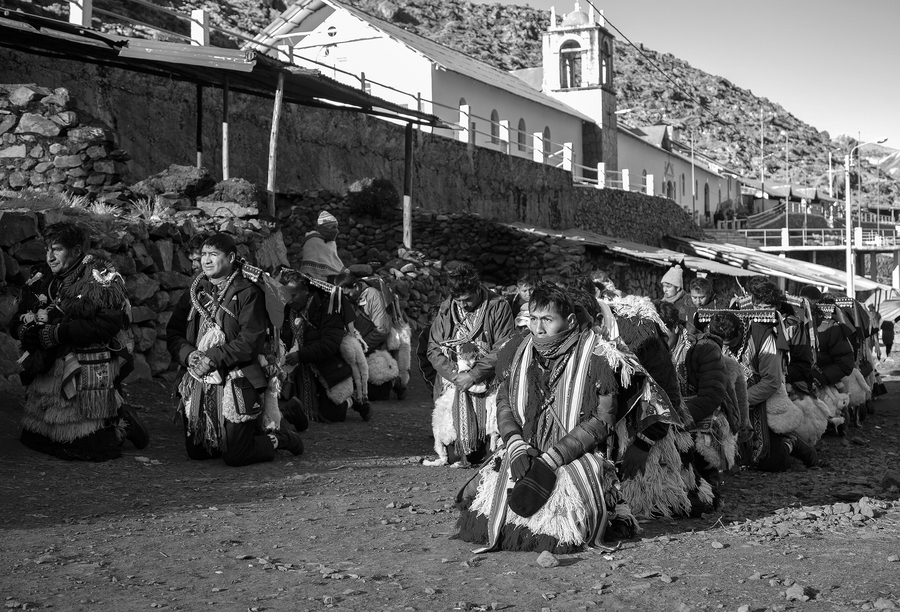 A black and white photo shows Peruvian men dressed in fringed costumes and woven scarves kneeling in lines on the ground outside a church.