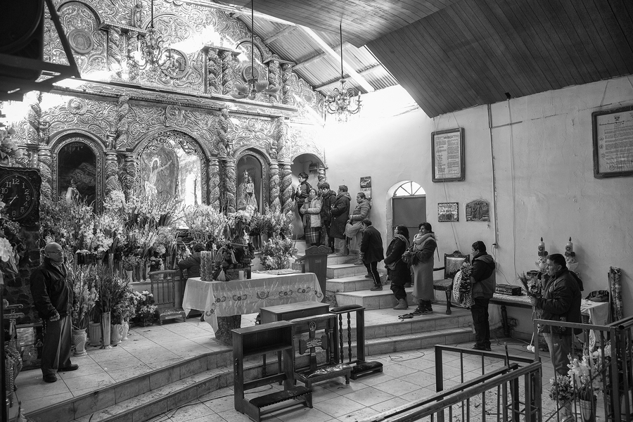 Inside a church, people wait in line to kneel and pray before an image of Christ as Lord of Qoyllur rit’i . Flowers decorate the altar and some of the pilgrims bring more flowers as offerings.