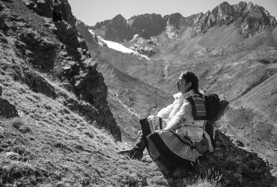 En una foto en blanco y negro, una peregrina peruana está sentada sobre una roca en un paisaje montañoso. Se veste una blusa con encaje y una falda rayada. Mira de manera pensativa hacía la montaña. 