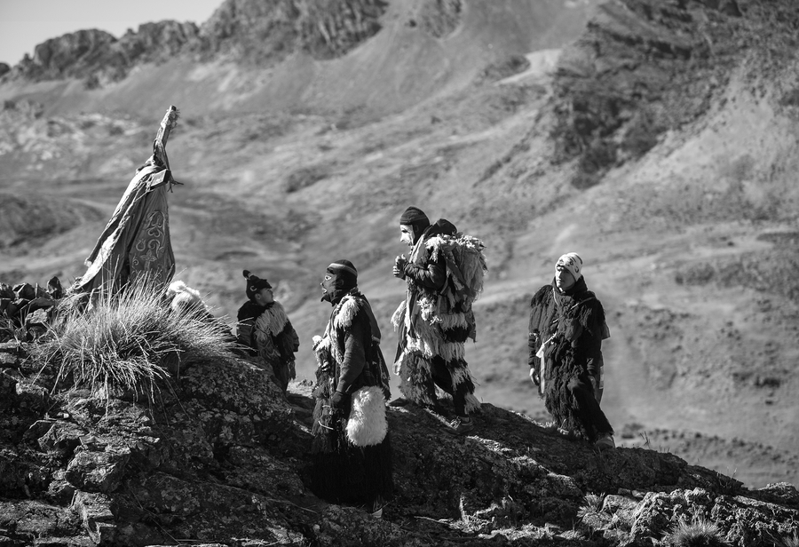 In a black and white photo, Peruvian men costumed in fringed tunics and ski masks approach a cross planted on a rocky mountain. The cross is dressed with a billowing cloth.