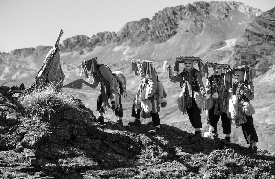 Un grupo de hombres vestidos en pasamontañas, bufandas, y sombreros con alas anchas y planas se juntan en una montaña. Se acercan a una cruz adornada con telas. 