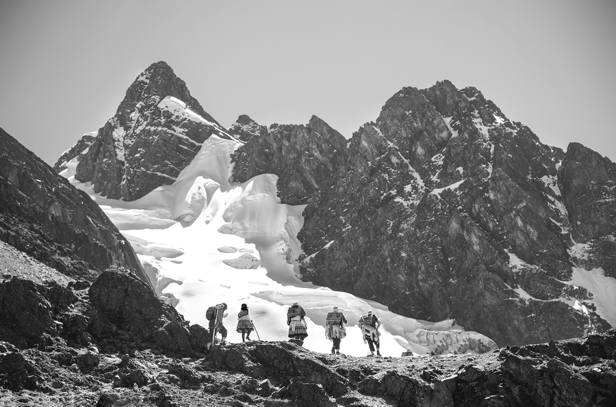 Photographed at a distance, five figures are visible climbing over a mountain range covered with snow. Their backs are to the camera, and they wear embroidered packs as they trek.