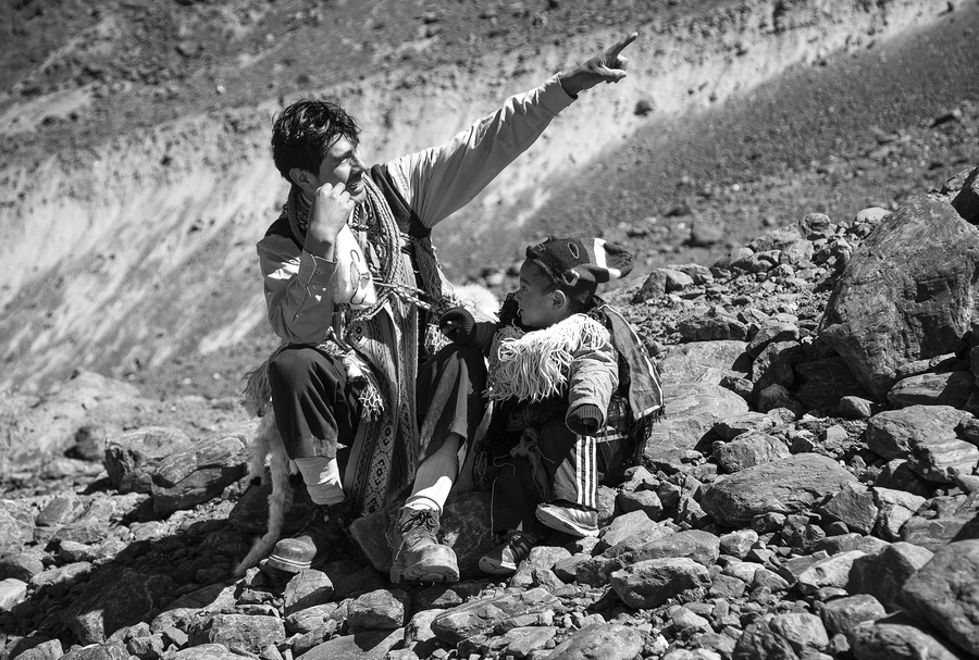 A black and white photo records a tender scene of a Peruvian father sitting on a craggy mountain with his young son. He points off into the distance up the mountain. They are bundled up in scarves, and the child wears a fuzzy hat.