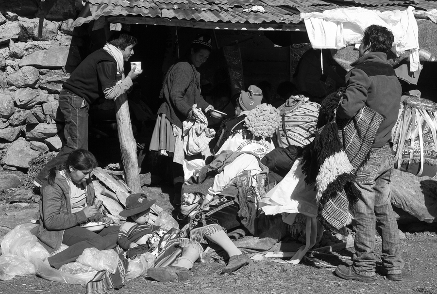 A black and white photo captures a group of pilgrims collapsed in exhaustion under a small, roofed stand. An older woman passes a plate to one of the gathered. A mother and small child sit off to the side eating.