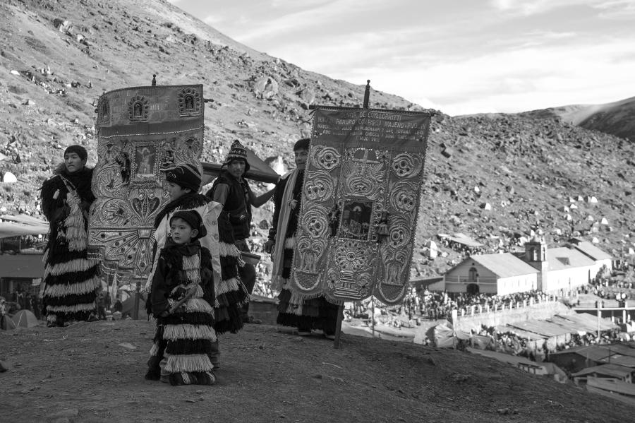 Un grupo de peregrinos peruanos de varias edades se juntan al aire libre. Se visten trajes con flecos y portan estandartes bordados con imágenes de Cristo, cruces, y llamas de fuego. 