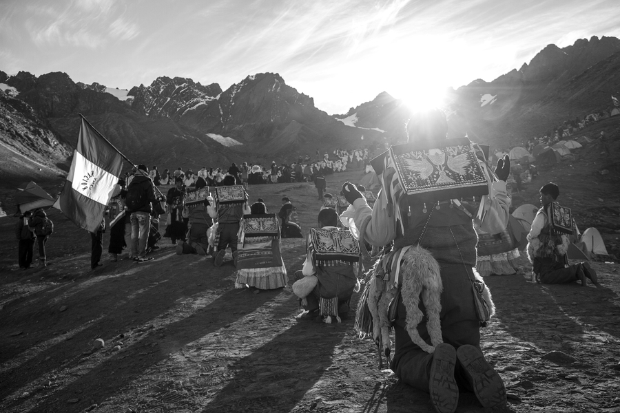 A black and white photo taken at daybreak captures pilgrims kneeling at the base of a mountain. They assemble in lines and are costumed in fringed or lacy outfits with embroidered hats on their backs. The sun shines through the mountain on the gathered.