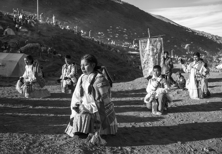 In a black and white photo, Peruvian pilgrims of different ages and genders kneel on the ground in prayer. They wear embroidered clothes and scarves. One pilgrim holds an embroidered banner standard.