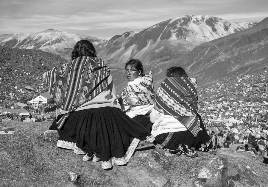 A black and white photo shows three Peruvian women sitting on a mountainside overlooking a large camp of pilgrims. Only one of the women turns around to face the camera. They all are dressed in skirts and woven shawls.