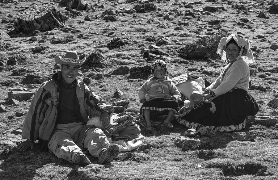 In a black and white photo, a Peruvian family sits outside on rocky ground. The father wears casual clothes, and the mother wears an embroidered skirt and a hat. Between them is a young girl in a Hello Kitty sweater and skirt. They smile at the camera.