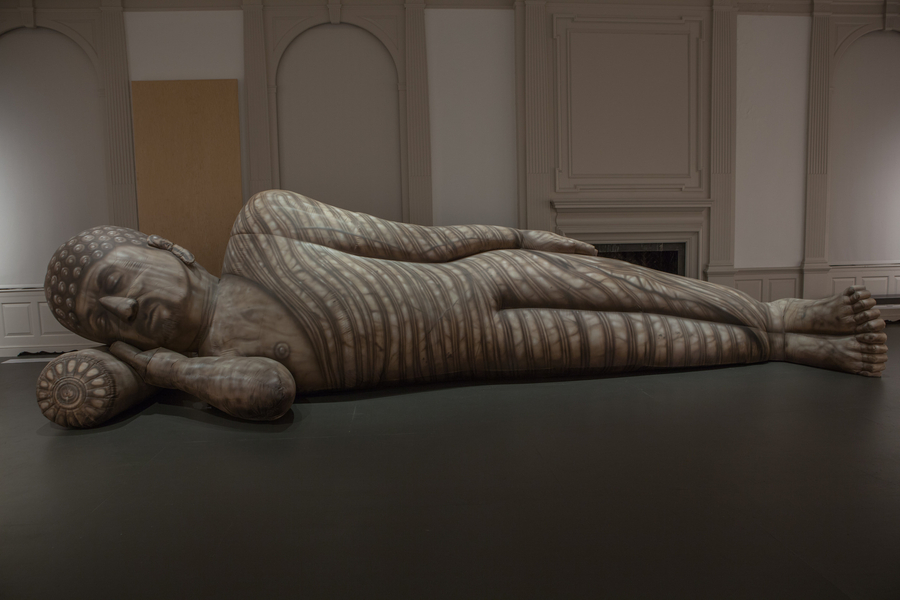 An enormous, inflated Buddha-like figure reclines on its side in a large room. He is made of a gray-colored cloth that is printed with all his facial and bodily features, including curly hair, an exposed nipple, and draping robes.