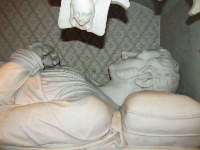 A detail of a large marble tomb effigy shows a man laid out with hands folded upon his chest and wearing a placid expression. The carved marble captures the drape of his clothes about him.