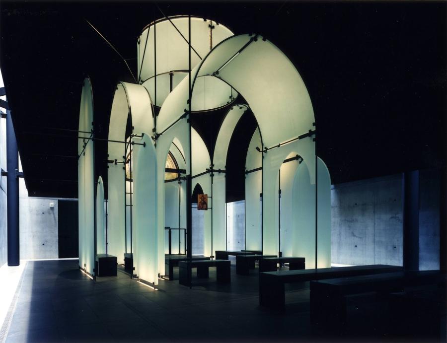 A freestanding architectural structure of suspended, opaque glass walls forms an abstracted basilica crossing and dome in a steel framework. The illuminated structure is filled with simple pews and enclosed in a large dark room.