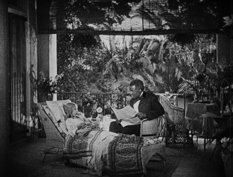A film still depicts a young, white girl lying in a rattan chaise beside a suited black man. The man holds a newspaper and reads to the girl. Tropical flora is visible over the open balcony in the background of their room.