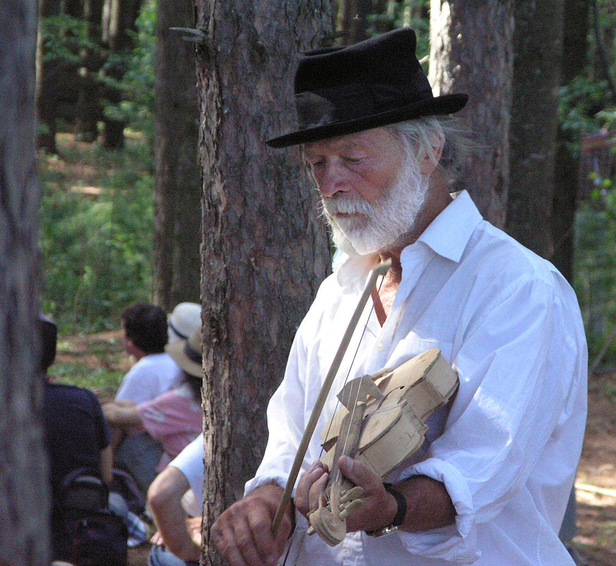 An older, light-skinned man plays a pale, wooden violin in a forest setting. He wears a black hat and looks down at his instrument. A group of people are visible in the background.