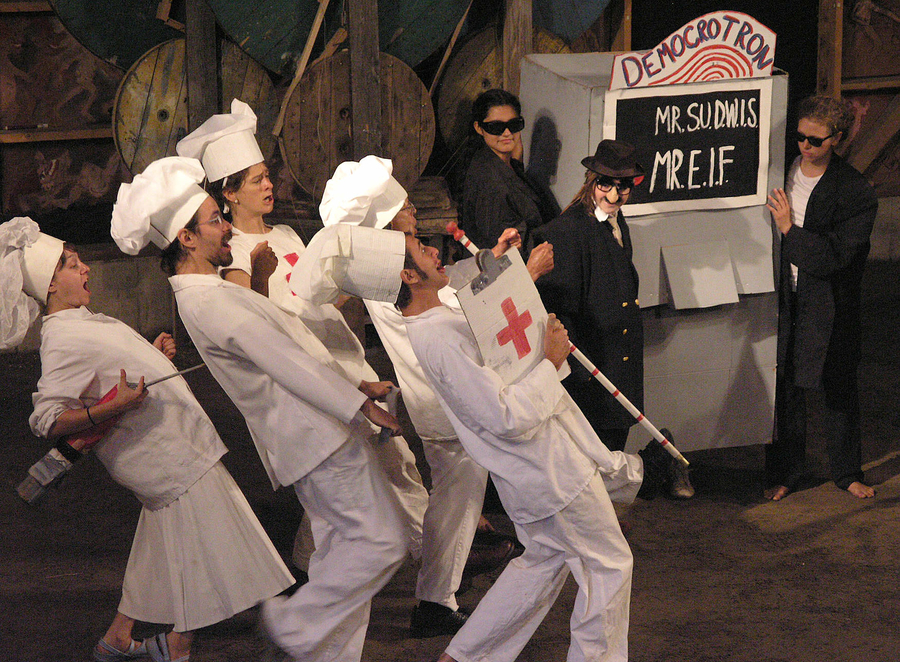 People dressed in white chef coats and hats approach a group dressed in black suits and sunglasses. They perform exaggerated movements as if in a play. 