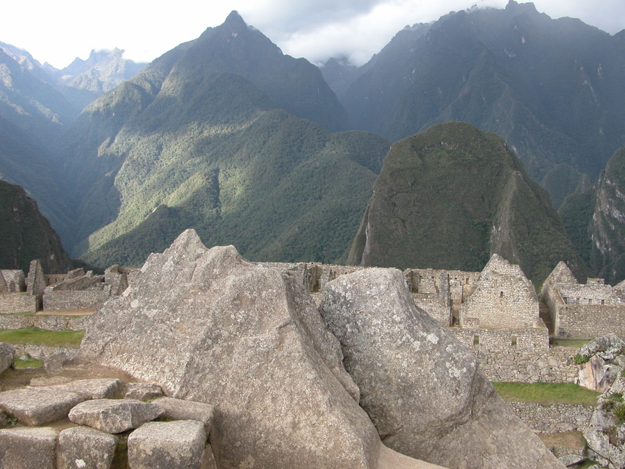 Gray, jagged stones stand in the foreground of a photo that captures a stone architectural ruins and green mountains in the background.