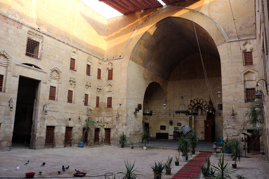 A view from within the courtyard of a khānqāh looks toward a massive pointed arch and low platform on one end. The wooden doors of khalwa cells line the ground floor of the building with the windows of living quarters up above looking onto the courtyard.