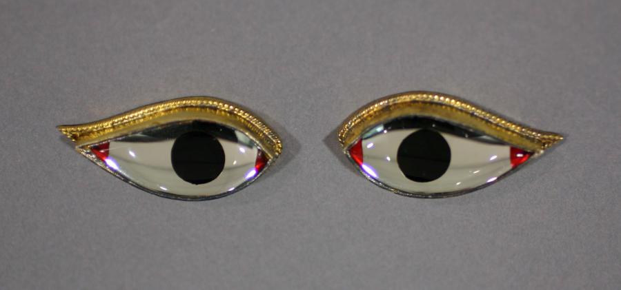 A pair of glass eyes with opaque, dark black irises have red paint at the corners and thin gold rims on top. Each eye has a slightly uplifted wing. 