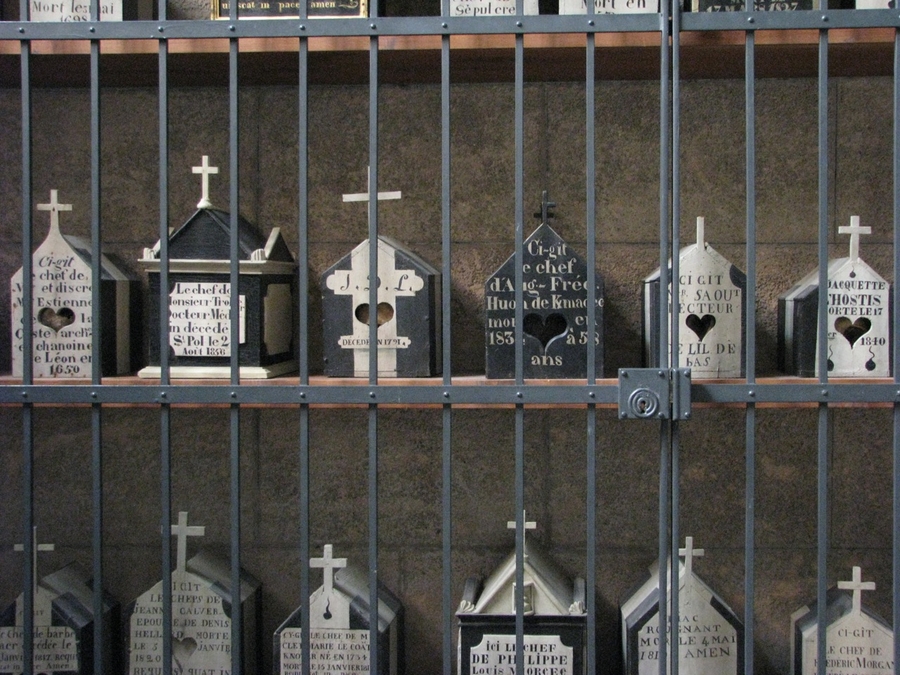 Three shelves of small gable-roofed and house-shaped boxes are displayed behind bars. Each box is black and white with a cross affixed to the top. All have text inscribed on the front.