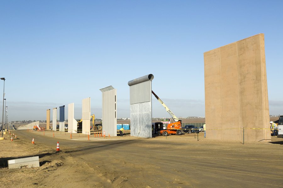 A row of tall panels are spaced out across a section of arid land. The panels are made of different materials but all the same shape and size. Construction vehicles and paraphernalia surround the panels.  