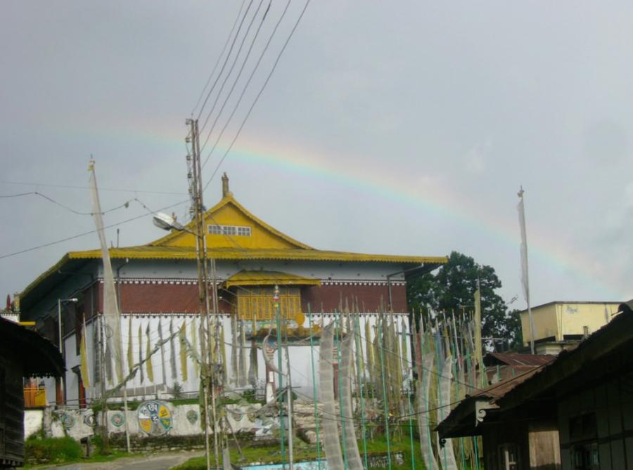 A rectangular monastery building has a flat roof with golden eaves and an ornamental gable. The monastery has a golden porch on its upper storey and a decorative red band encircling the structure. Flags are set up outside the monastery.