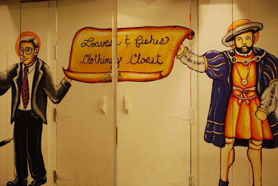 A haloed, tan-skinned man in a black suit and a haloed tan-skinned man in regal 16th century clothes are painted on either side of white closet doors. They hold a yellow banner painted over the doors that reads "Loaves and Fishes Clothing Closet."