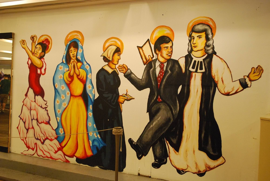 A section of a mural of dancing, haloed figures depicts a tan-skinned woman in a red flamenco dress and Our Lady of Guadalupe. They are among other figures including a man in a suit kicking up his feet and clasping his arm around a light-skinned cleric.