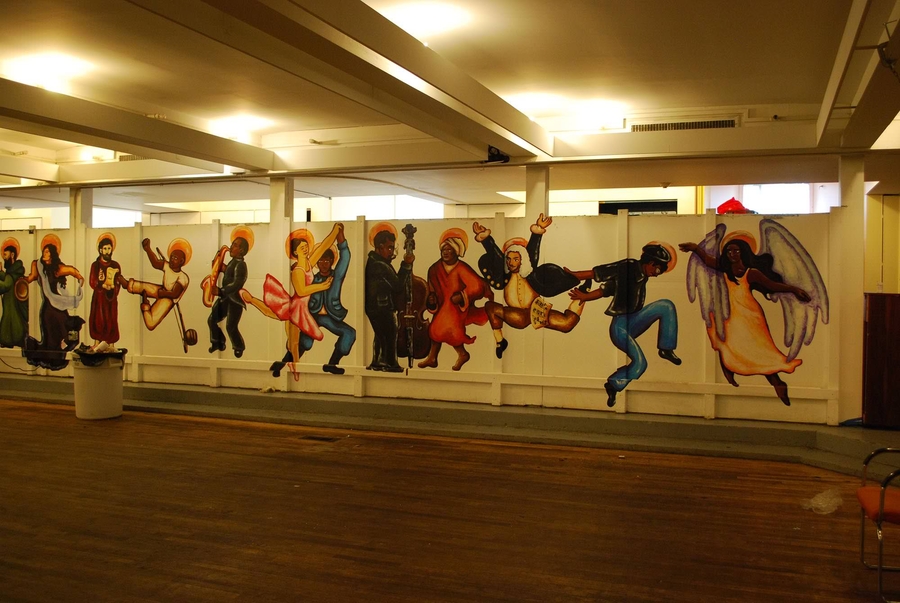 A photo of a church basement room shows a mural of dancing figures from different time periods leaping and playing music on a long horizontal wall.