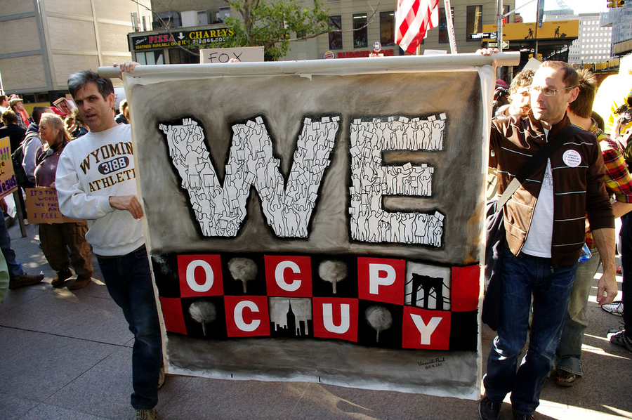 Two light-skinned men hold up a banner at a protest. It reads, "WE OCCUPY." The letters of WE are filled with sillhouettes of protesters. The letters of OCCUPY are checkered and interspersed with photos of Manhattan and nuclear explosions. 