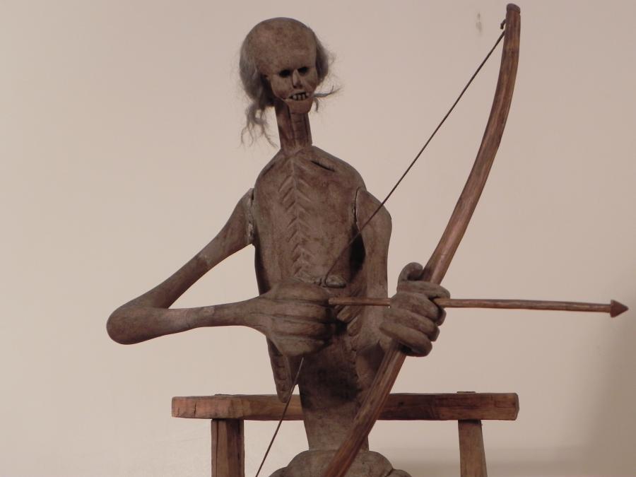 A wooden sculpture depicts a skeletal figure pulling a bow and arrow taunt. He has a flat chest imprinted with ribs, a head of wispy gray hair and large teeth, and oversized hands.