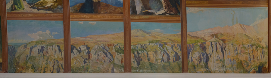 A church mural depicts a craggy mountain landscape overlooking blue waters beneath a bright blue sky. Wooden ribbing runs over the scene.