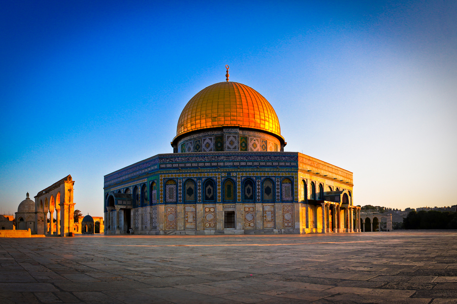 A photo captures the Dome of the Rock against a bright blue sky. The Islamic shrine consists of a golden dome atop a large octagonal base coated with blue and green mosaics.