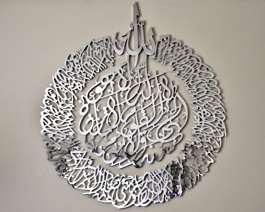 A dense piece of Arabic calligraphy carved out of stainless steel wraps around an embellished center. The text is a portion of the Qur’an and the work is crowned by the word "Allah." 