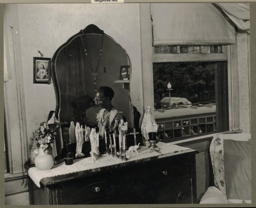 In a black and white photo, a mirrored dresser is set with chalkware statues of the Virgin and saints alongside candles and crucifixes. A dark-skinned woman is visible sitting before the dresser in the mirror. 