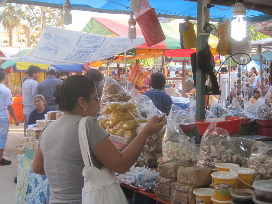 A tan-skinned woman examines the nuts, fruits, and treats assembled on a stall in a crowded market. 