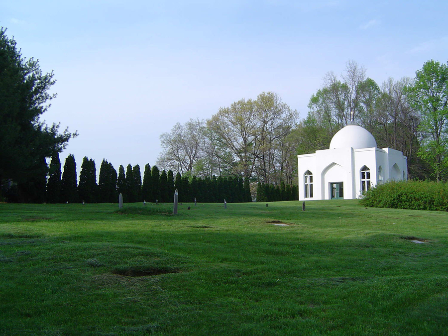 A green grassy lawn scattered with short wooden posts lies in front of a white shrine. There is a border of hedges and other greenery on the far left side.