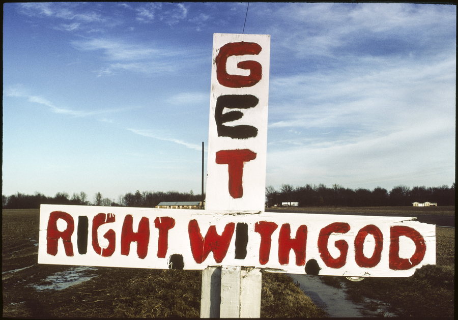 A white sign is shaped like a cross and painted with red text. The vertical axis reads "GET." The horizontal axis reads, "RIGHT WITH GOD."