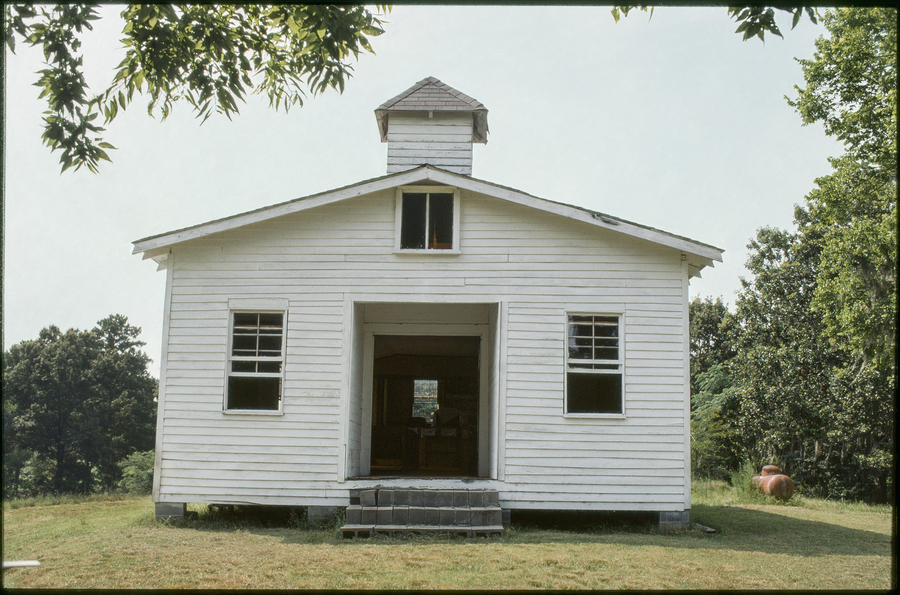 A white church looks like a small house with wooden siding. No doors close off the entrance so the viewer can look through the building to the window at the far end. One sees out to the trees behind the photographed church.