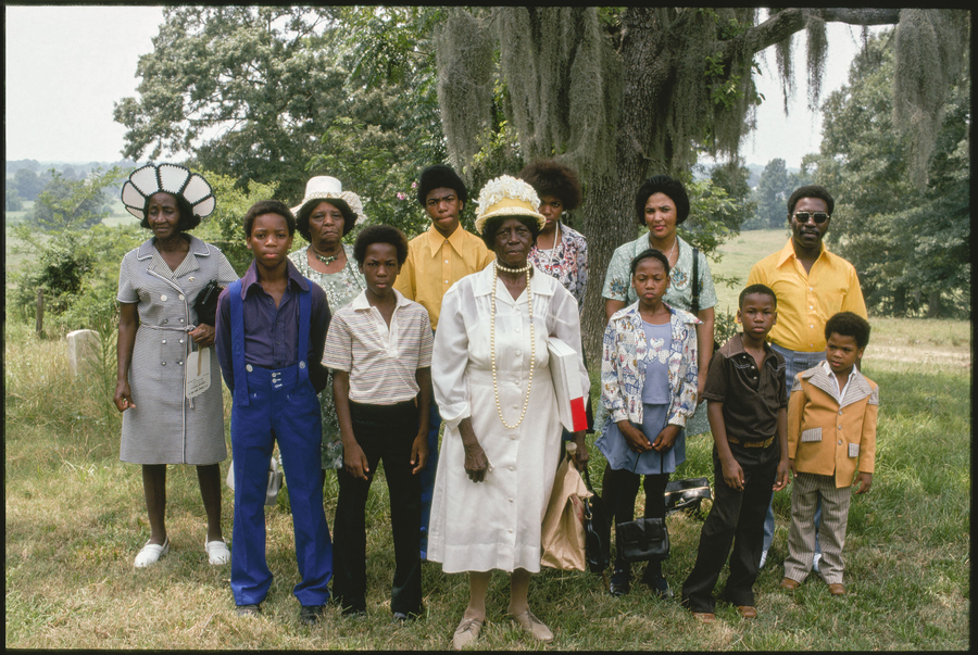 A small group of black churchgoers assemble in front of a large green tree. An elderly woman in a crisp white dress, large pearls, and hat stands at the center of the group. The others vary in age and gender. All face the camera with a solemn expression.